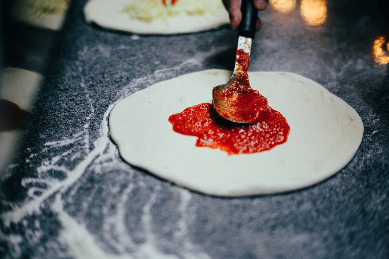 Spreading pizza sauce over pizza dough with the back of a spoon