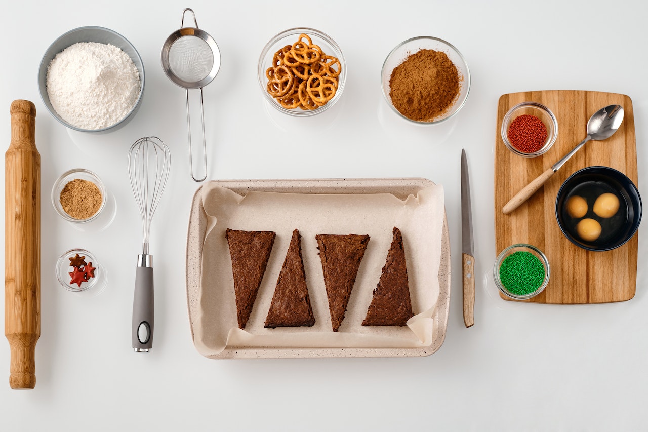 A table with different baking ingredients, including a rolling pin, a bowl with flour, a whisk, a tray with cake slices, a cutting board with additional ingredients, a bowl with pretzels, etc.