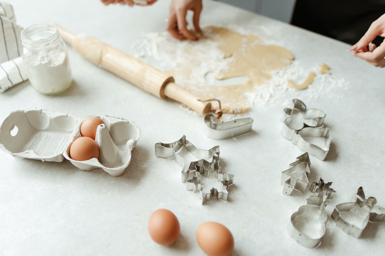 A person spreading out cookie dough on flour, eggs, a rolling pin, and cookie cutters on the table
